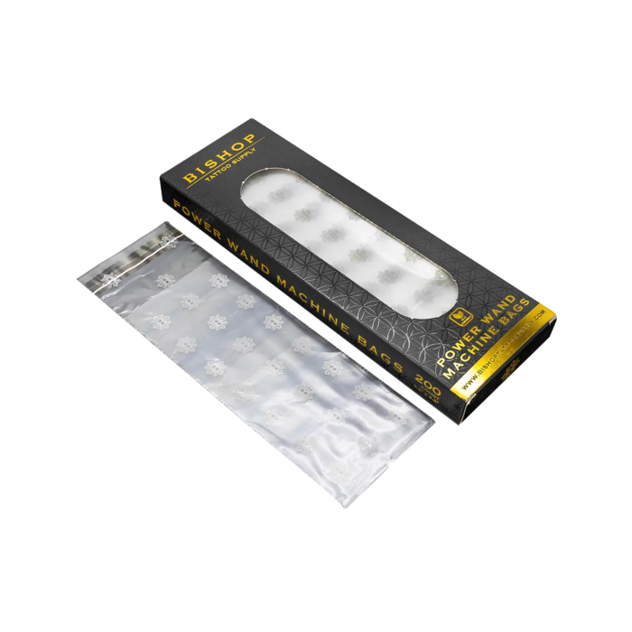 Une boîte rectangulaire noire et dorée étiquetée Sacs pour Machine Bishop Power Wand, avec une fenêtre transparente, à côté d’une pile de sacs en plastique transparents ornés de motifs de flocons de neige blancs.